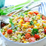 A bowl of macaroni pasta salad with chopped tomatoes, bell peppers, celery, red onion, and parsley, with serving utensils on top.