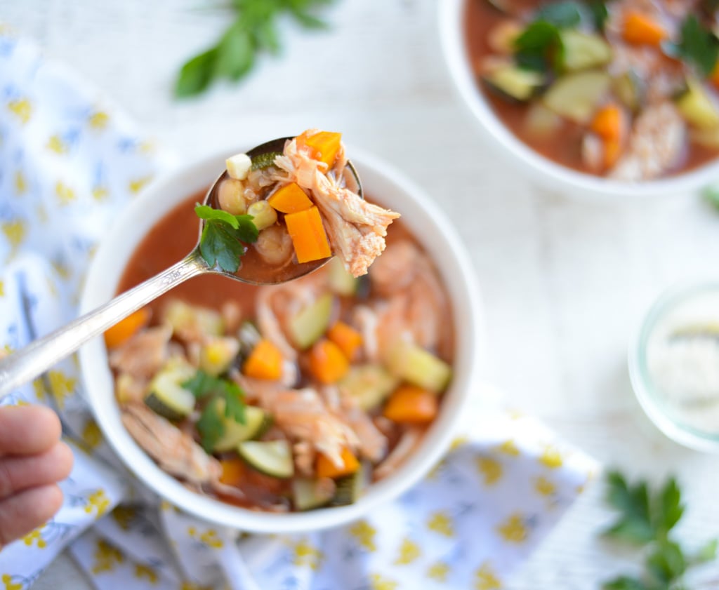 A spoonful of chicken and vegetable soup held above a bowl, with visible carrots, zucchini, and herbs on a light surface.