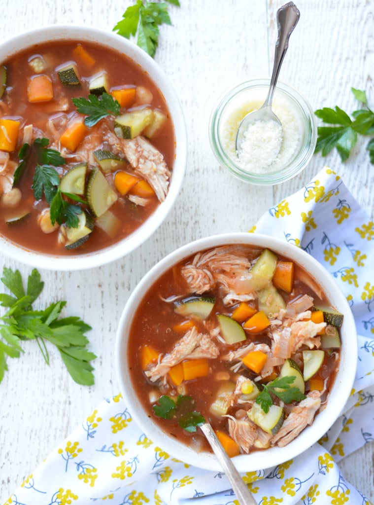 Two bowls of vegetable and chicken soup with tomato broth are on a white surface, garnished with parsley. A small jar of grated cheese with a spoon and a floral napkin are nearby.