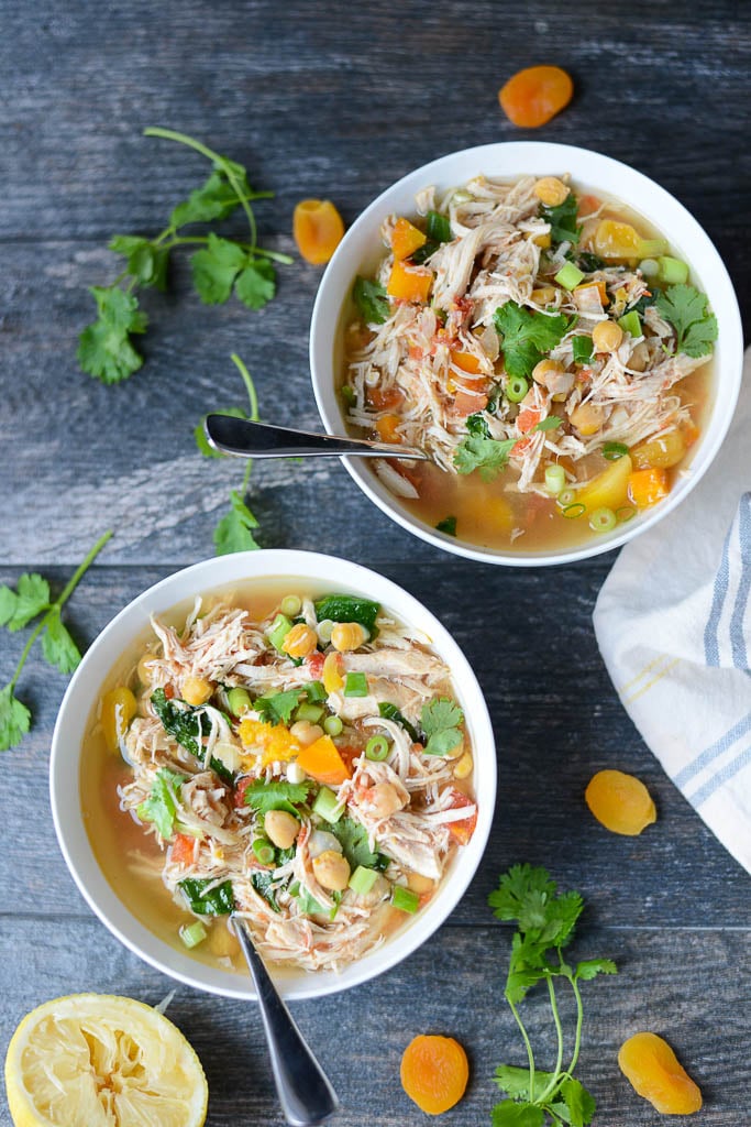 Two bowls of shredded chicken soup with vegetables and herbs, garnished with sliced green onions and cilantro, on a wooden surface with dried apricots and lemon.