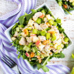 Close up of a chicken apple salad in a white square bowl, with a red apple in the background.