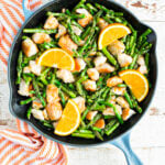 Blue and black pan with orange chicken asparagus stir-fry and orange slices on top with a striped orange napkin on a wood table.