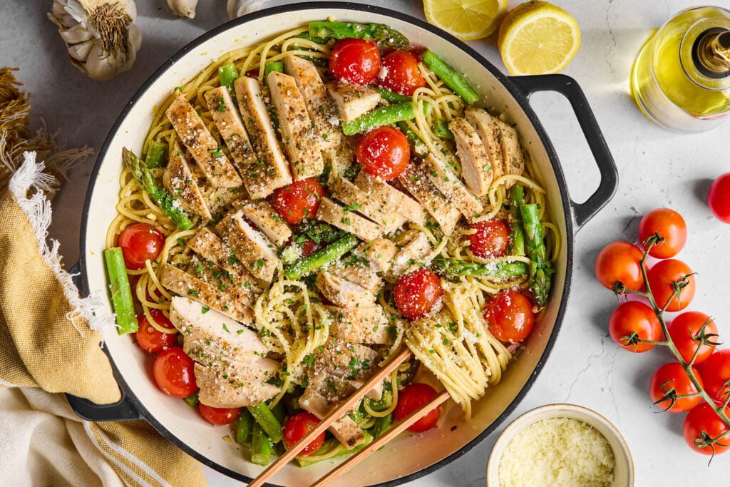 A pan of spaghetti with sliced grilled chicken, cherry tomatoes, asparagus, and grated cheese, surrounded by fresh tomatoes, lemon halves, garlic, and olive oil.
