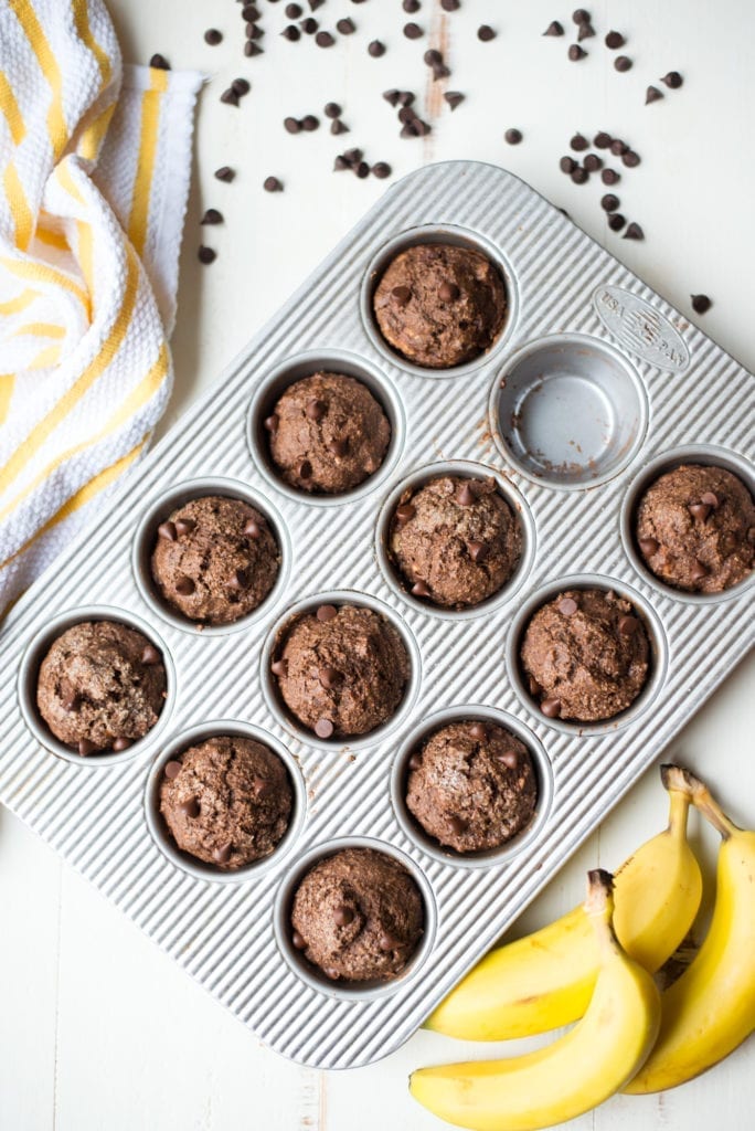 A metal muffin tin holds eleven chocolate muffins with one empty slot, surrounded by chocolate chips, a yellow-striped towel, and two bananas on a white surface.