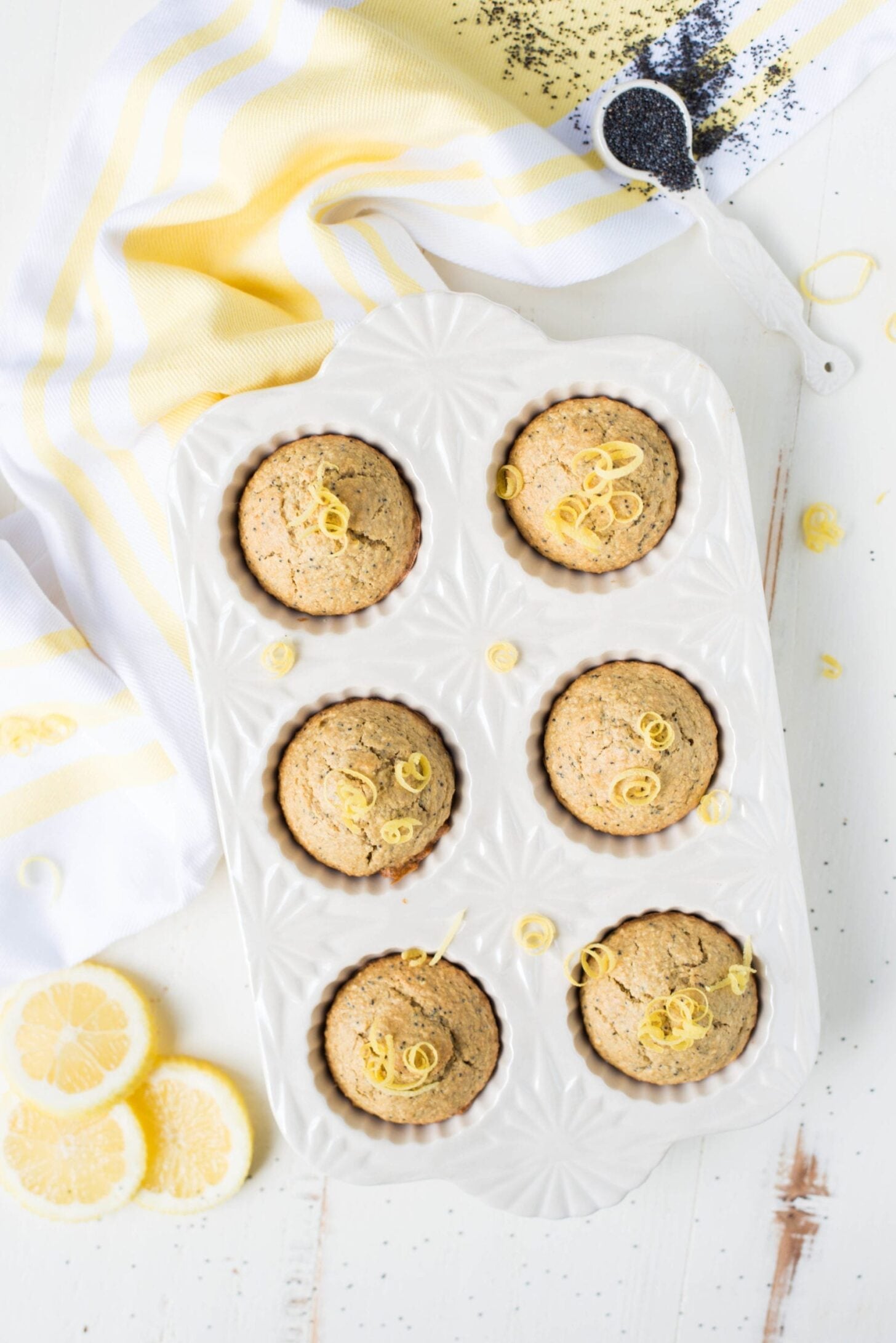 A white muffin tray holds six lemon poppy seed muffins, topped with lemon zest. Sliced lemons, a striped towel, and a spoon of poppy seeds are on the white surface nearby.