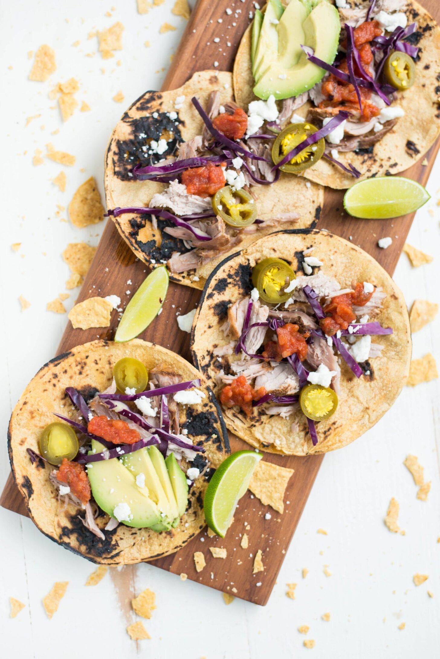 Four grilled corn tortillas topped with shredded cabbage, avocado slices, feta cheese, jalape&ntilde;os, salsa, and lime wedges, arranged on a wooden board with tortilla crumbs scattered around.