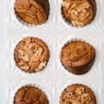 Six baked muffins, some topped with walnuts, arranged in a white ceramic muffin tray with a geometric pattern.