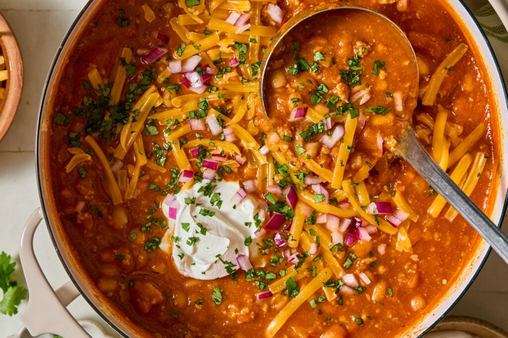 A pot of chili topped with shredded cheddar cheese, chopped red onions, chopped cilantro, and a dollop of sour cream, with a ladle resting inside.