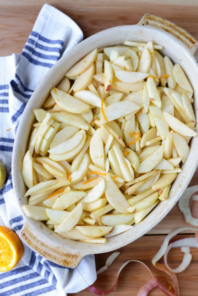 Oval baking dish filled with peeled and sliced apples, placed on a striped kitchen towel with apple peels and an orange on the wooden surface nearby.