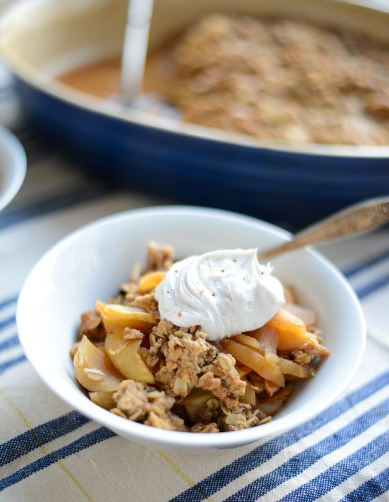 A bowl of apple crisp topped with a dollop of whipped cream, with a spoon, sits on a striped cloth; a baking dish of apple crisp is in the background.
