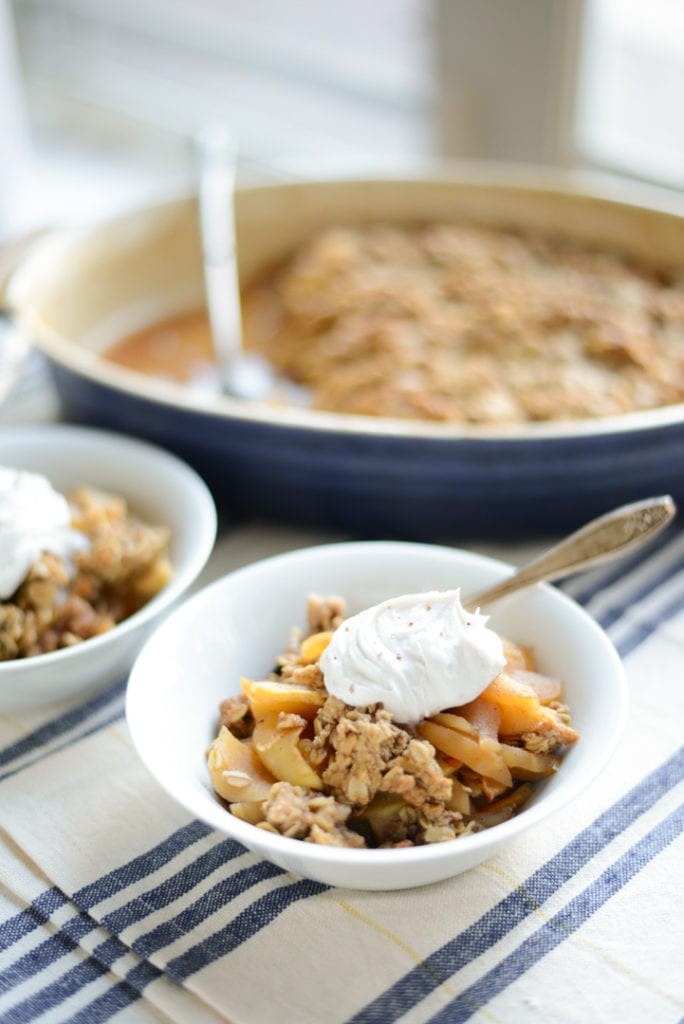 Two bowls of apple crisp topped with whipped cream sit on a striped cloth, with a larger dish of apple crisp in the background.