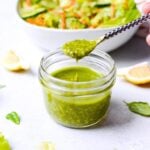 spoon dipping into a small glass jar of pesto vinaigrette with salad in the background