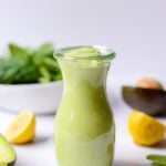 glass jar of green salad dressing with sliced lemons and cut avocados on a white background