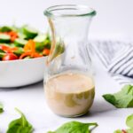 side view of creamy balsamic dressing in a clear glass carafe surrounded by leafy greens on a white table