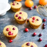 Six cranberry muffins in gold wrappers are arranged on a wooden surface with loose cranberries, white baking cups, orange fruits, and a whisk in the background.