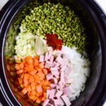 Chopped ham and split peas with onions and carrot in a black crockpot on white background.