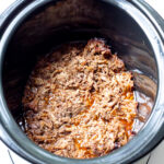 Shredded cooked meat sits at the bottom of a black slow cooker with visible juices.