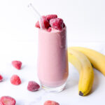 strawberry banana peanut butter smoothie in a clear glass surrounded by frozen fruit on a white background