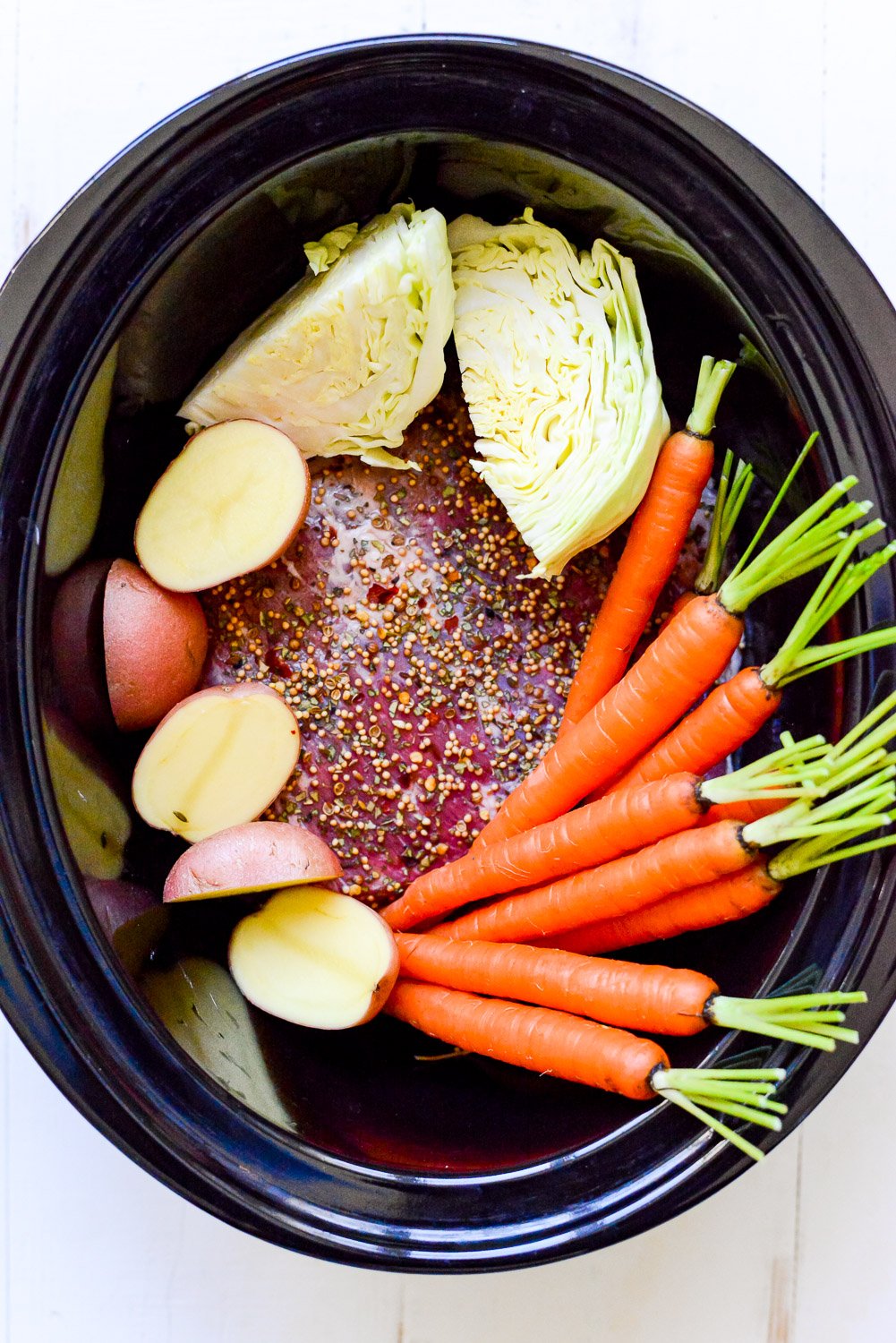 top down view of slow cooker corned beef ingredients in a black crockpot on a white background