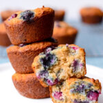 A stack of three blueberry muffins sits on a plate, with one muffin split open in front to reveal a moist, berry-filled interior. More muffins are visible in the background.