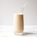 Clear glass with a clear straw filled with an oat milk smoothie on a white background surrounded by rolled oats.