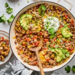 A skillet filled with a ground turkey, beans, corn, and ground meat dish, topped with melted cheese, avocado slices, sour cream, cilantro, and chopped green onions. A wooden spoon rests inside.