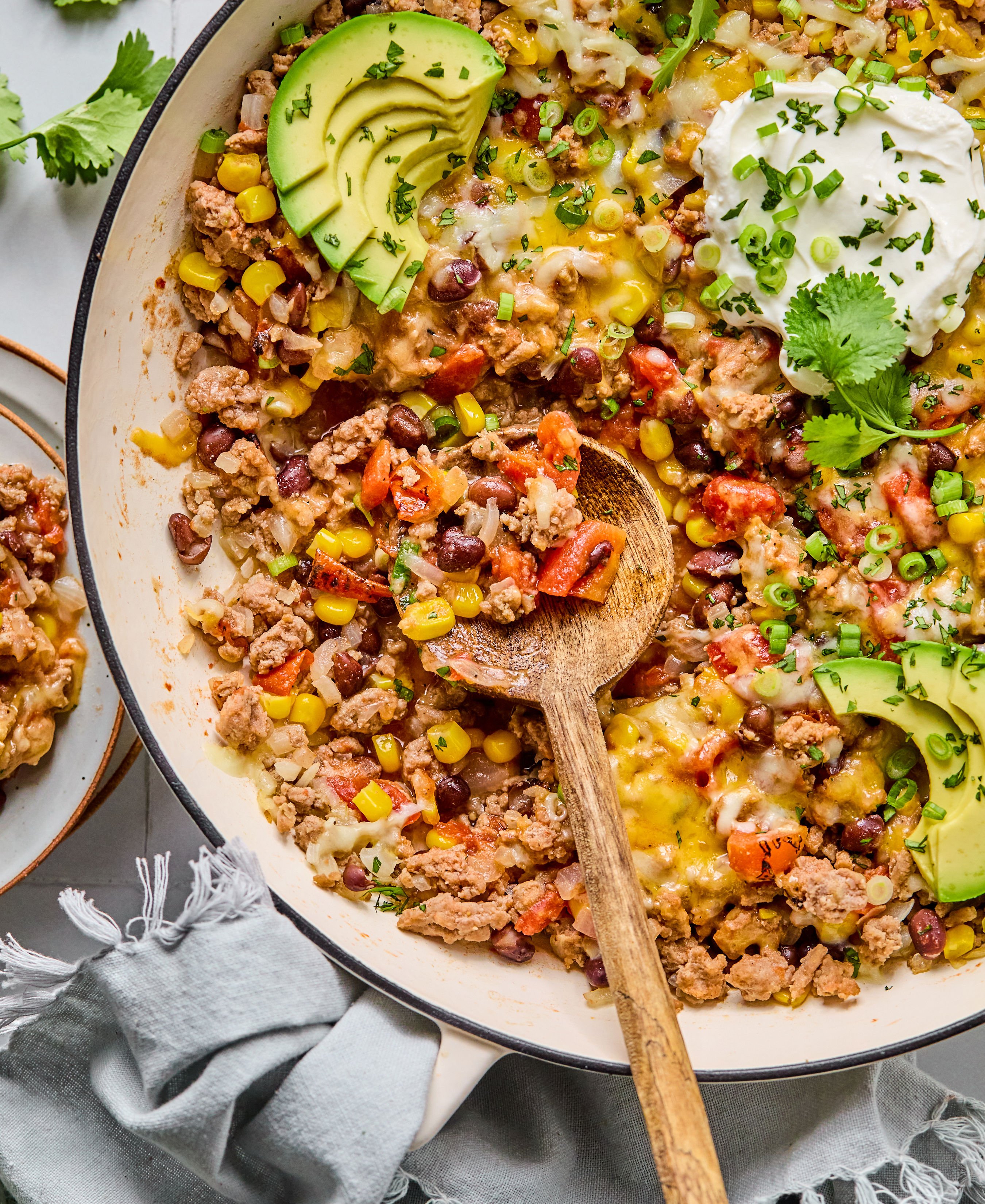 A wooden spoon in a skillet filled with ground turkey, chopped vegetables, and sliced avocado. 