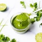 Cilantro lime sauce being spooned out of a glass jar, with fresh herbs and lime wedges alongside it.