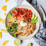 White dish filled with instant pot chicken burrito bowl filling with avocado slices on top, sitting on a white background with a napkin and chips on the side.