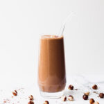 A clear glass filled with a nutella smoothie with a straw, surrounded by hazelnuts on a marbled background.