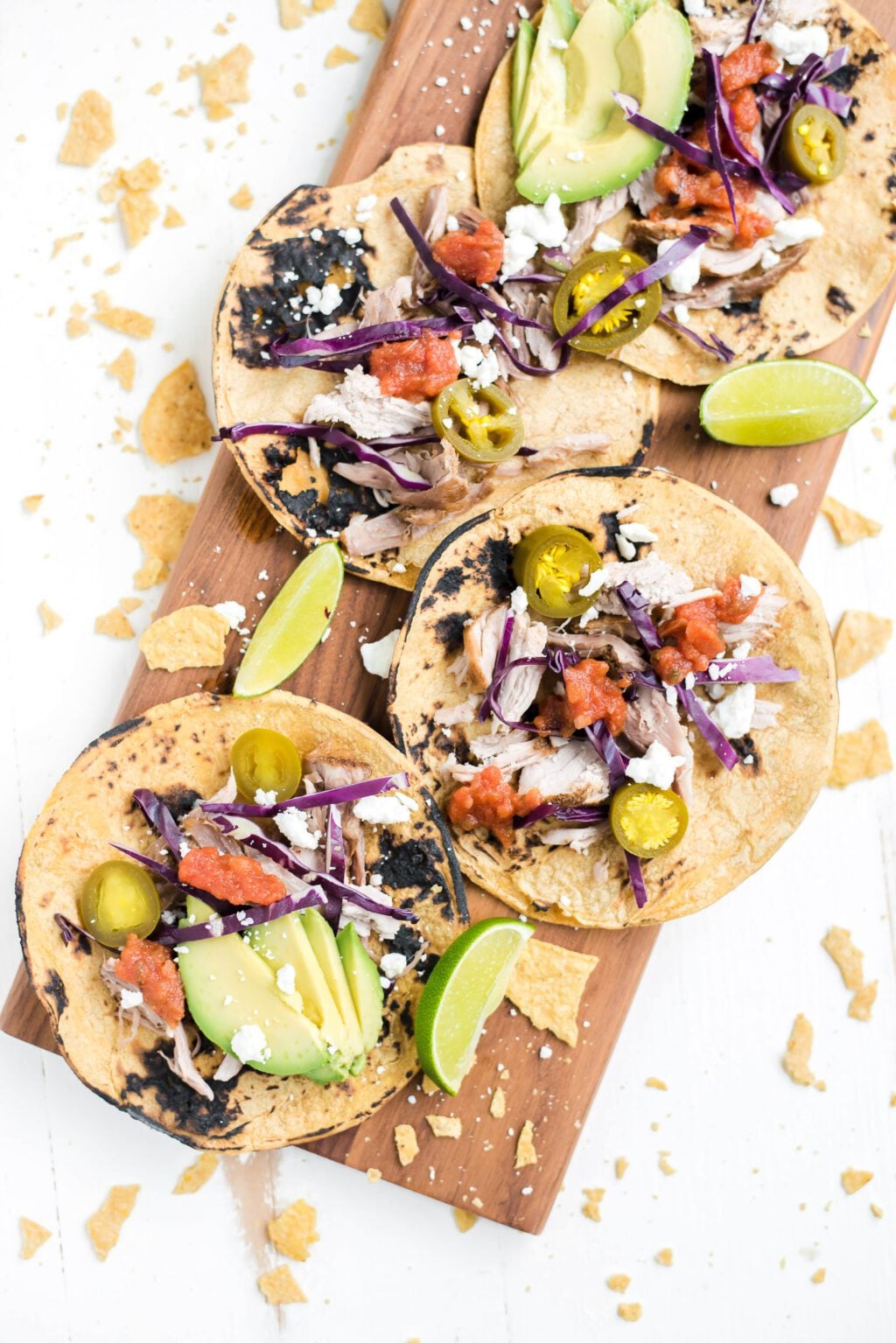 Three grilled corn tortillas topped with shredded chicken, avocado, jalape&ntilde;os, cabbage, salsa, feta cheese, and lime wedges on a wooden board. Tortilla chips are scattered around.