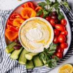 Top down view of a vegetable platter with a bowl of yogurt tahini sauce in the center, on a white background with a napkin on the side.