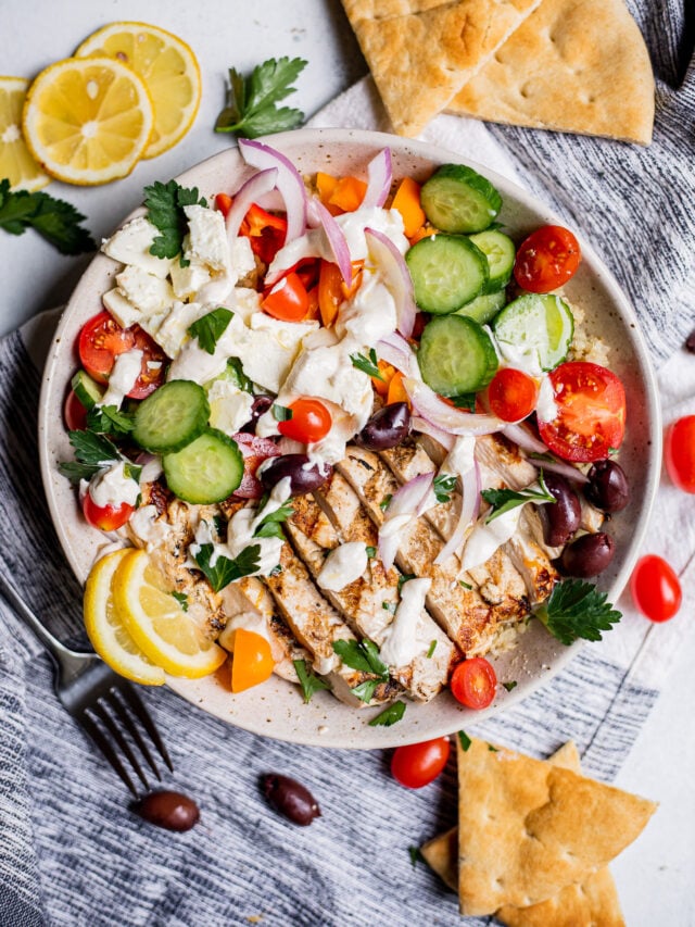 A bowl of Greek salad with grilled chicken, feta, cucumbers, tomatoes, olives, red onions, parsley, lemon slices, pita bread, and dressing, on a white surface.