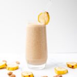 Clear glass filled with a banana almond milk smoothie surrounded by banana slices on a white marbled background.