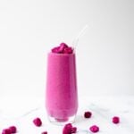 A clear glass filled with a dragon fruit smoothie surrounded by frozen fruit pieces, on a white marbled background.