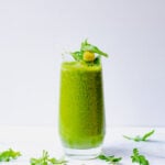 Side shot of a bright green arugula smoothie with pineapple on top and arugula strewn around the bottom.