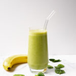 Shot of a green banana spinach smoothie in a glass jar with glass straw and banana and baby spinach.
