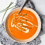 Top down view of a white bowl filled with butternut squash and red pepper soup, with a striped napkin on the side.