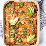 Overhead shot of a baking pan filled with chicken enchiladas with fresh avocado slices and jalapenos on top.