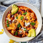 Brown bowl on a blue background filled with chicken and bean chili, topped with cheese, cilantro, and avocado.