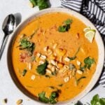 Overhead shot of a white bowl filled with sweet potato peanut soup, topped with fresh herbs, on top of a white background.