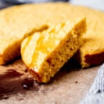 A close-up of a sliced piece of cornbread drizzled with honey, resting on a wooden surface with a napkin nearby.