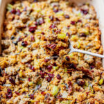 A baking dish filled with cornbread stuffing containing celery and dried cranberries, with a serving spoon lifting a portion.