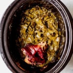 Overhead shot of cooked collard greens with a ham hock in a black slow cooker.