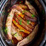 Overhead shot of slow cooker flank steak topped with fresh herbs inside of a black crockpot.