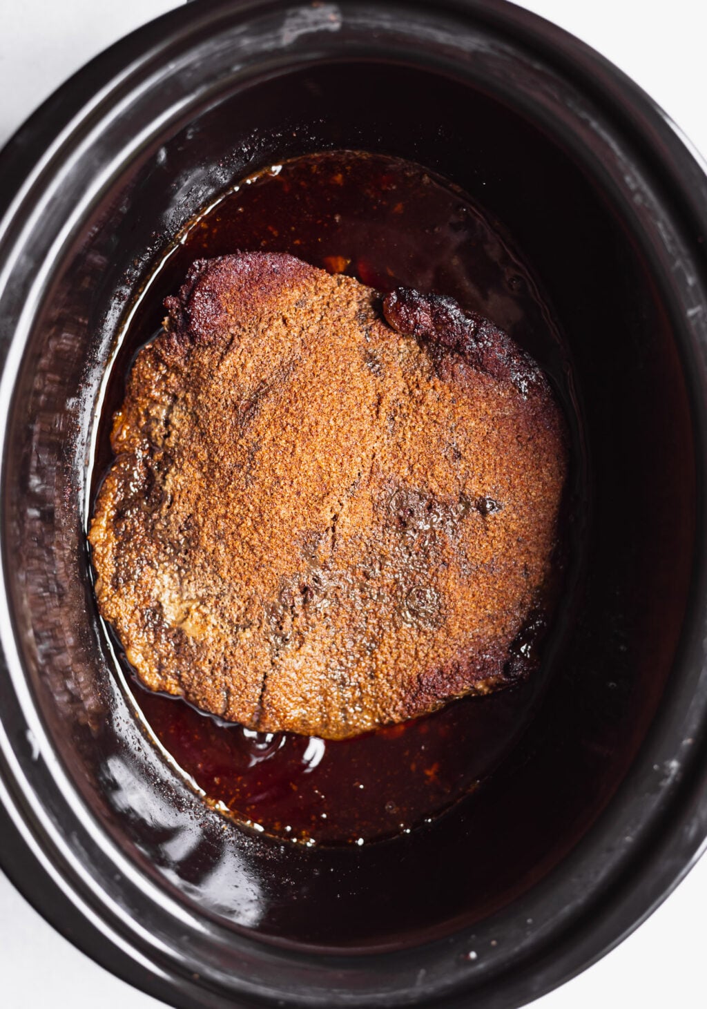 A seasoned piece of meat is slow-cooking in a black slow cooker with visible juices surrounding it.