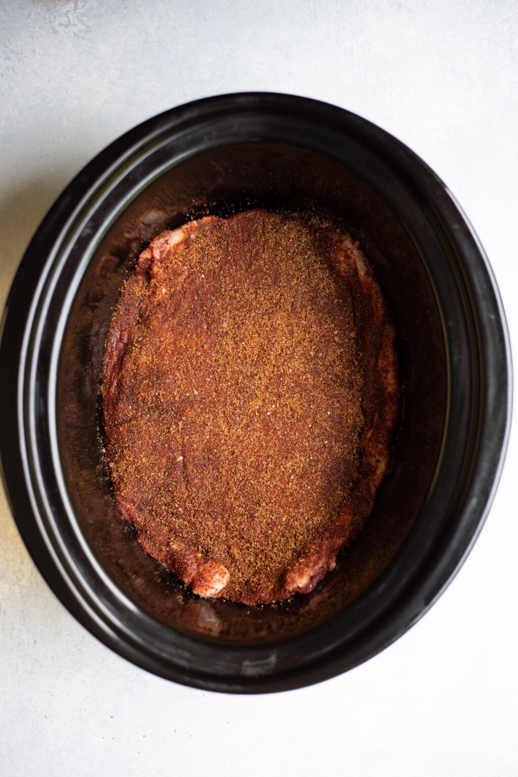 Raw meat covered in a layer of dry seasoning sits at the bottom of a black slow cooker.