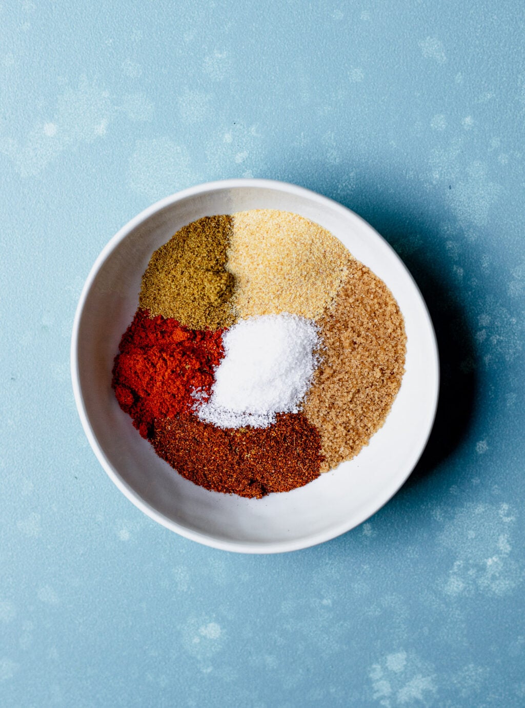 A white bowl containing six different spices and seasonings, arranged in separate sections, on a blue textured surface.