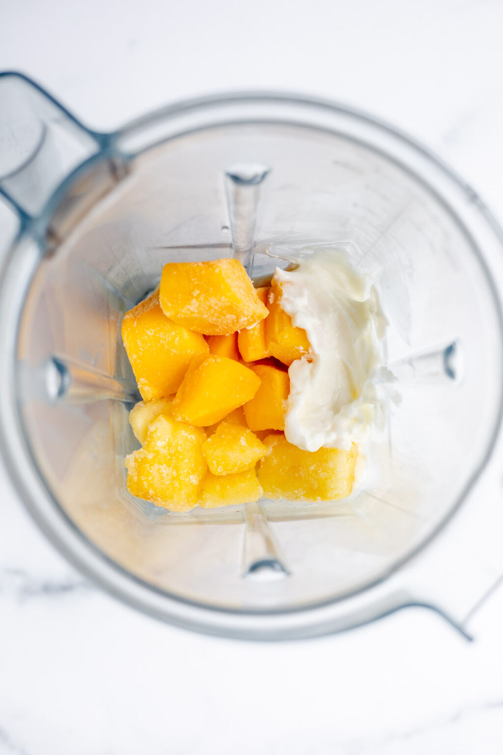 Overhead view of a blender containing frozen mango chunks and a dollop of yogurt on a white surface.