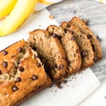Side shot of sliced banana bread with chocolate chips on a cutting board with bananas in the background.